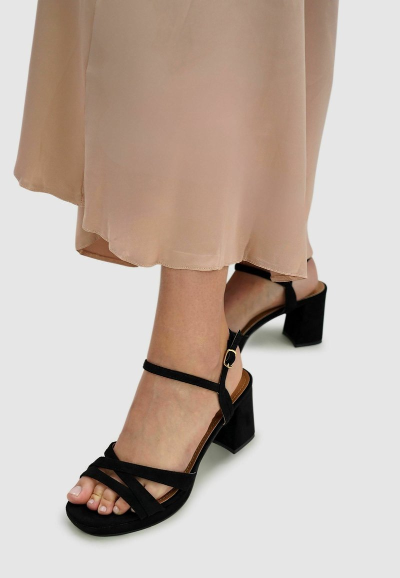Black suede heeled sandals featuring a multi-strap design, ankle straps with a gold buckle, and a textured block heel.