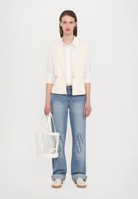 Gilet crema chiaro sopra una camicia bianca, abbinato a jeans azzurri chiari con un design stampato e una borsa trasparente bianca.