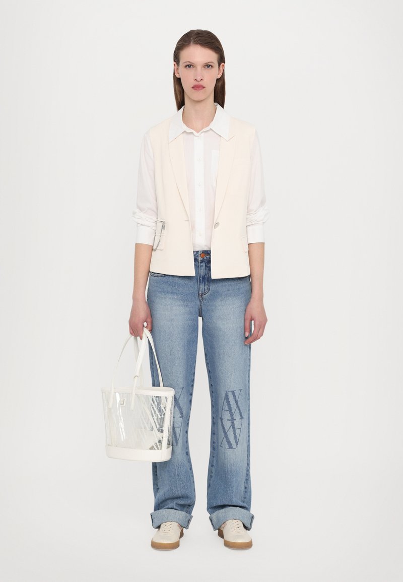 Gilet crema chiaro sopra una camicia bianca, abbinato a jeans azzurri chiari con un design stampato e una borsa trasparente bianca.