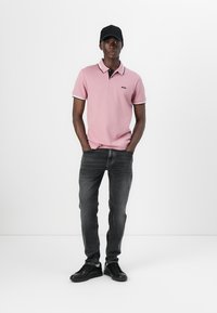 Rosa pikétröja med kontrasterande svart krage, grå jeans och svarta sneakers; modellen står med händerna i fickorna mot en vit bakgrund.