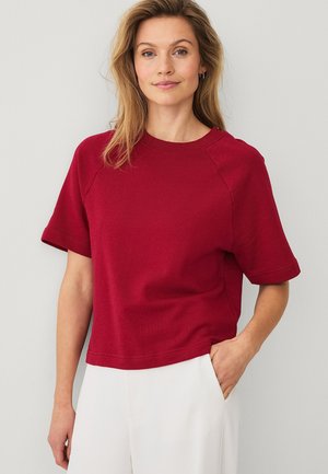 Kvinde iført en rød kortærmet sweater og hvide bukser, stående med den ene hånd i lommen mod en ensfarvet lys baggrund.