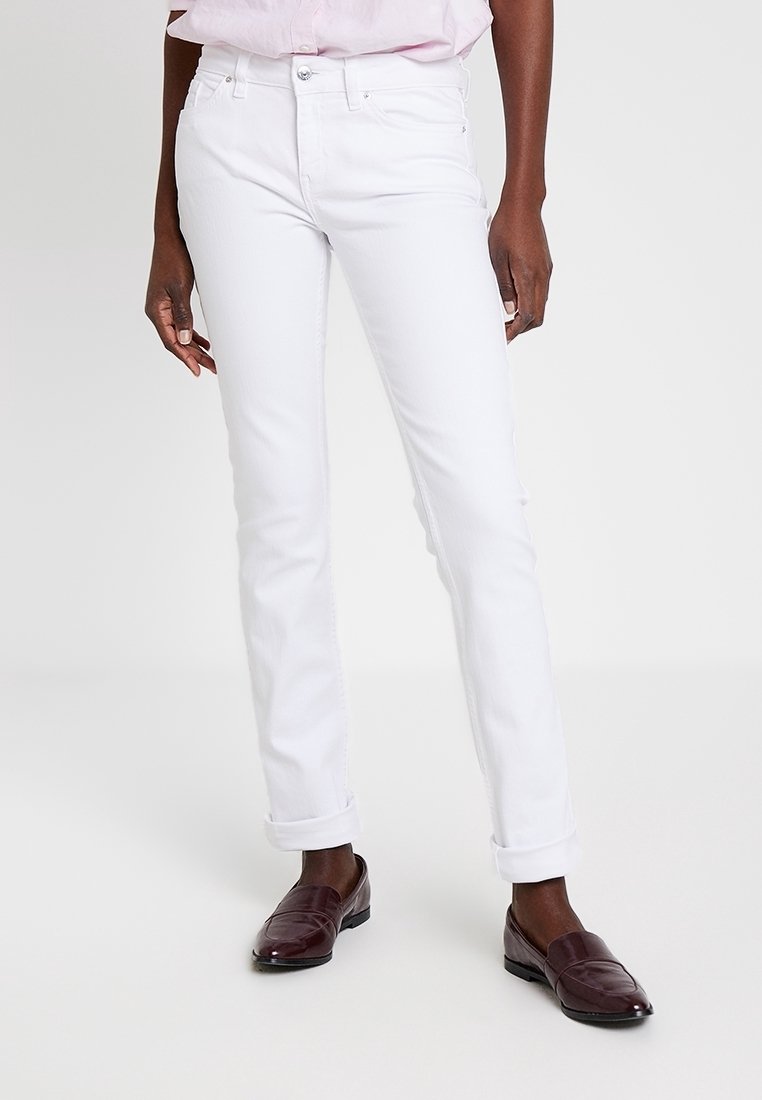 Person trägt weiße, schmal geschnittene Jeans mit Umschlag, hellrosa Hemd und dunkelbraune Loafers, steht vor einem schlichten hellen Hintergrund.