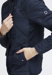 Námornícky prešívaný kabát s diamantovým vzorom, s otvorením na zips, bočnými vreckami a detailom loga na hornej rukáve. Materiál s jemnou textúrou.