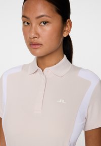 Młoda kobieta stojąca przodem, ubrana w jasnobeżową koszulkę polo z białymi akcentami na ramionach oraz małym logo na piersi.