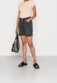 Černé denimové šortky s vysokým pasem, spárované s světle béžovým pruhovaným tričkem a černými sandály na klínu. Drží vzorovanou černobílou tašku.