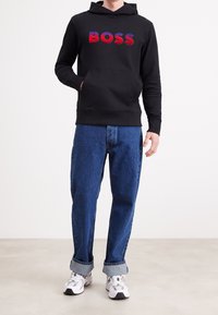 Felpa nera con cappuccio con la scritta "BOSS" in lettere rosse e blu, abbinata a jeans di denim blu e sneakers bianche. Vestibilità casual, trama semplice.