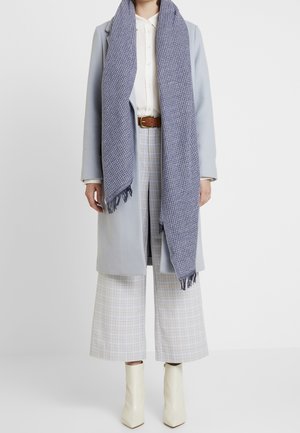 Femme portant un manteau bleu clair, une écharpe bleue à motifs avec franges, un chemisier blanc, un pantalon large à carreaux, une ceinture marron et des bottines blanches.