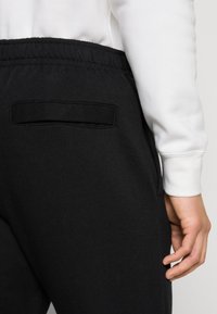 Pantalones de chándal negros con cintura elástica y un bolsillo trasero. La tela parece suave con una textura lisa, combinados con una sudadera blanca.