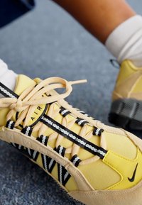 Sapatilhas amarelas com materiais de malha e camurça, apresentando atacadores pretos e brancos, e uma sola texturizada. Rótulo "AIR" na lateral.