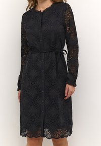 Spetsklänning i marinblått med långa ärmar, knappknäppning fram och midjeband. Har blommönster och vågig fåll. Andningsaktiv textur.