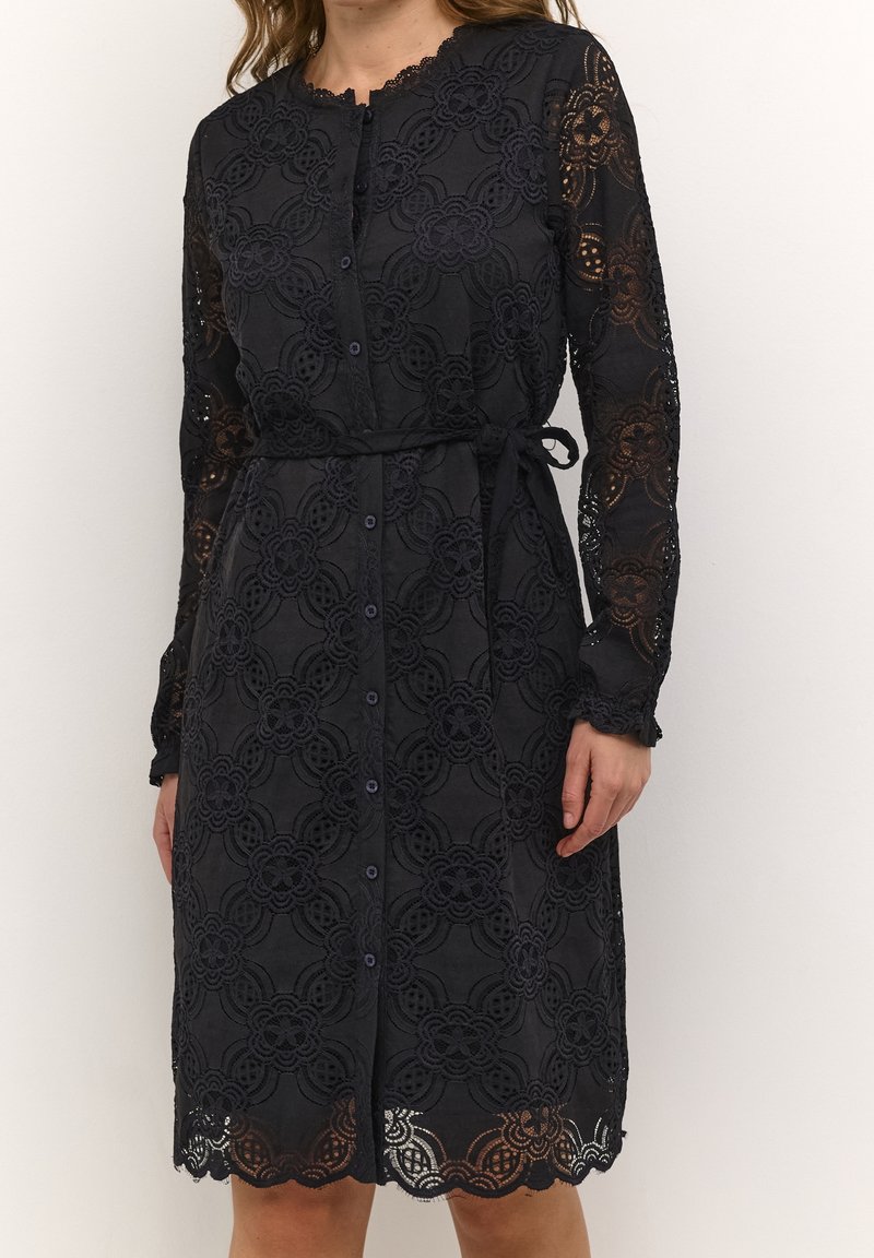 Spetsklänning i marinblått med långa ärmar, knappknäppning fram och midjeband. Har blommönster och vågig fåll. Andningsaktiv textur.