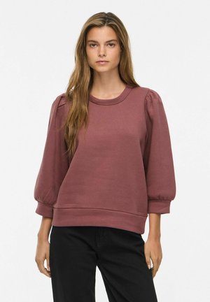 Frau mit langen Haaren, die einen mausfarbenen Pullover mit Puffärmeln und schwarze Hosen trägt, steht vor einem einfarbigen hellen Hintergrund.