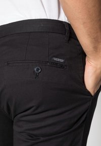 Pantaloni neri con vestibilità slim, realizzati in un tessuto liscio. Presentano una tasca posteriore con chiusura a bottone e un'etichetta con il marchio vicino alla vita.