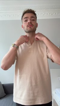 Top in maglia beige a maniche corte e scollo a V, con una leggerezza di trama, vestibilità rilassata e piccolo logo sul petto. Indossato con le mani sul colletto.