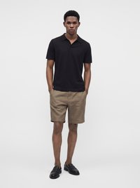 Mann steht frontal, Hände in den Taschen, trägt ein schwarzes Poloshirt, beigefarbene Shorts und schwarze Loafer vor einem weißen, schlichten Hintergrund.
