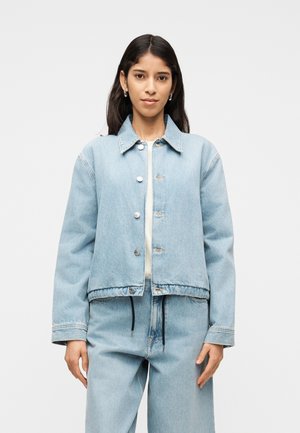 Žena s dugom tamnom kosom, odjevena u svijetloplavu traper jaknu i odgovarajuće traper hlače visokog struka, na jednoličnoj pozadini.