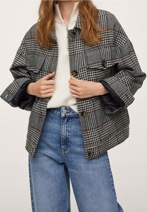 Persona que lleva una chaqueta de cuadros en blanco y negro con botones sobre un suéter blanco con cremallera, combinados con unos vaqueros de talle alto azul.