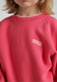 Różowa bluza z miękkiego materiału z prążkowanym dekoltem i mankietami. Z logo „AMERICAN VINTAGE” w białym kolorze. Prosty design, luźny krój.