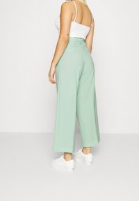 Pantalons larges vert menthe à taille haute, avec une texture lisse et des poches arrière, accompagnés de baskets blanches.