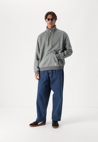 Sudadera gris con cremallera y cuello alto, bolsillo frontal y dobladillo acanalado; combinada con jeans de mezclilla azul holgados y zapatillas negras.