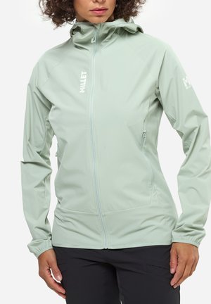 Femme portant une veste à capuche zippée vert clair avec poches latérales zippées et un pantalon noir, debout sur un fond blanc.