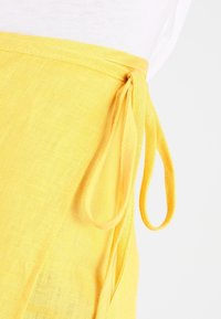 Gelbes Leinenkleidungsstück mit einer Bindeschließe an der Seite. Der Stoff hat eine strukturierte Oberfläche und ein leichtes, atmungsaktives Design.