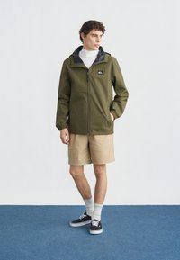 Νέος άνδρας όρθιος, φοράει χακί ζακέτα με φερμουάρ, μπεζ σορτς, άσπρες κάλτσες και μαύρα αθλητικά παπούτσια σε μπλε πάτωμα με λευκό φόντο.