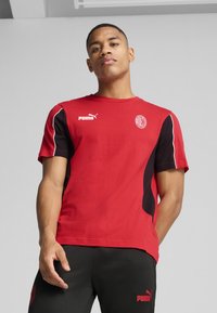 Červené tričko s krátkym rukávom s čiernymi akcentmi a bielym prešívaním. Obsahuje logo Puma na ľavej hrudi a kruhový emblém na pravej strane.