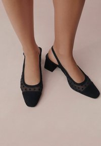 Musta slingback-kenkä, jossa on neliönmuotoinen kärki, mokkanahkainen materiaali, pistekuviollinen verkkopäällys ja leveä korko. Kenkaassa on läpinäkyvä pilkkukuosi.