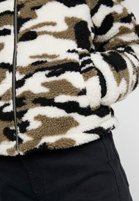 Luddig, mönstrad fleecejacka med ett kamouflage-mönster i beige, svart och vitt. Har dragkedja fram och sidofickor.