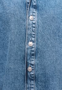 Gros plan sur un tissu en denim bleu clair avec une rangée verticale de boutons-pression argentés au centre.