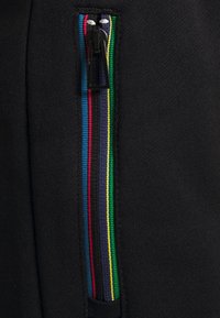 Schwarze Hosen mit einem Reißverschluss und einem mehrfarbigen gestreiften Band auf der Seite, die Textur und moderne Designdetails zeigen.