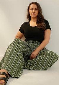 Svart åtsittande T-shirt, grön- och vitrandiga vida byxor med knytmidja, sittande pose, slät textur och minimal metall.