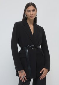 Black tailored blazer with a single button closure and long sleeves. Features a black leather belt and sleek design. Smooth texture.