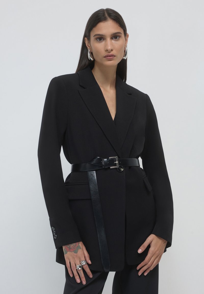 Blazer nero su misura con chiusura a un bottone e maniche lunghe. Include una cintura in pelle nera e un design elegante. Texture liscia.