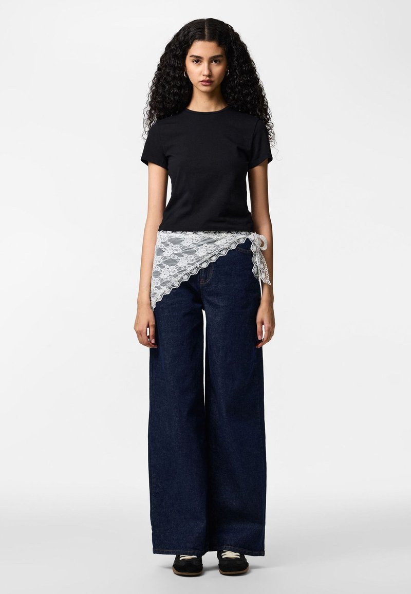 Giovane donna con lunghi capelli ricci indossa una maglietta nera, jeans scuri a gamba larga, scarpe nere e una cintura di pizzo bianca annodata in vita.