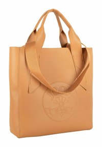 Syntetická taška přes rameno v barvě koňaku s textura povrchu, má dva prodloužené uchy a vpředu reliéfní logo. Čtvercový tvar.