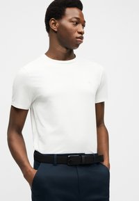 Νεαρός άνδρας με απλό λευκό κοντομάνικο πουκάμισο, φορεμένο μέσα σε σκούρο μπλε παντελόνι με μαύρη πλεκτή ζώνη, με τα χέρια στις τσέπες, κοιτάζοντας προς το πλάι.