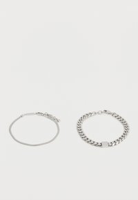 Δύο βραχιόλια σε ασημί τόνο σε λευκό φόντο: το ένα έχει λεπτή αλυσίδα με ορθογώνια και εξαγωνικά γούρια, το άλλο έχει χοντρή αλυσίδα τύπου curb με τετράγωνο γούρι.