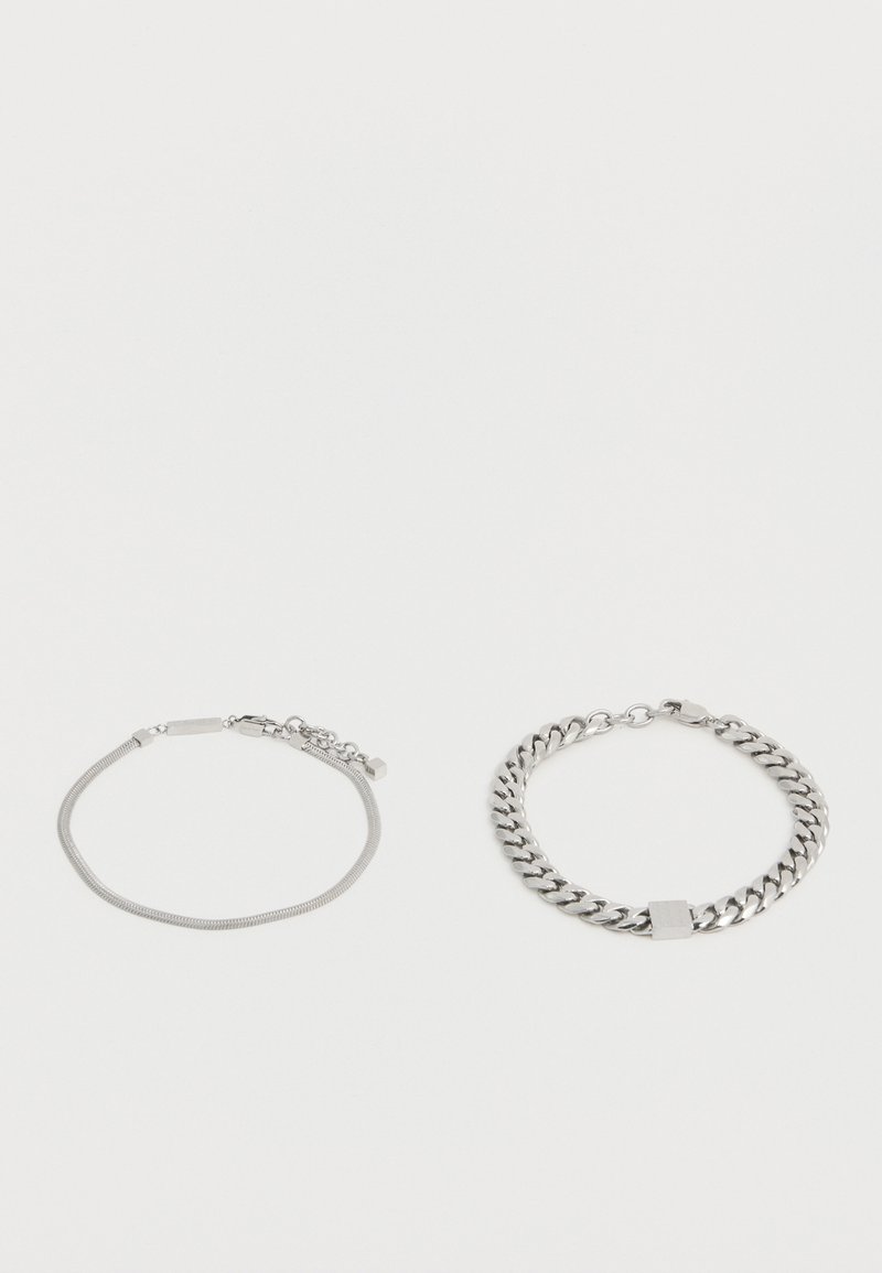 Δύο βραχιόλια σε ασημί τόνο σε λευκό φόντο: το ένα έχει λεπτή αλυσίδα με ορθογώνια και εξαγωνικά γούρια, το άλλο έχει χοντρή αλυσίδα τύπου curb με τετράγωνο γούρι.