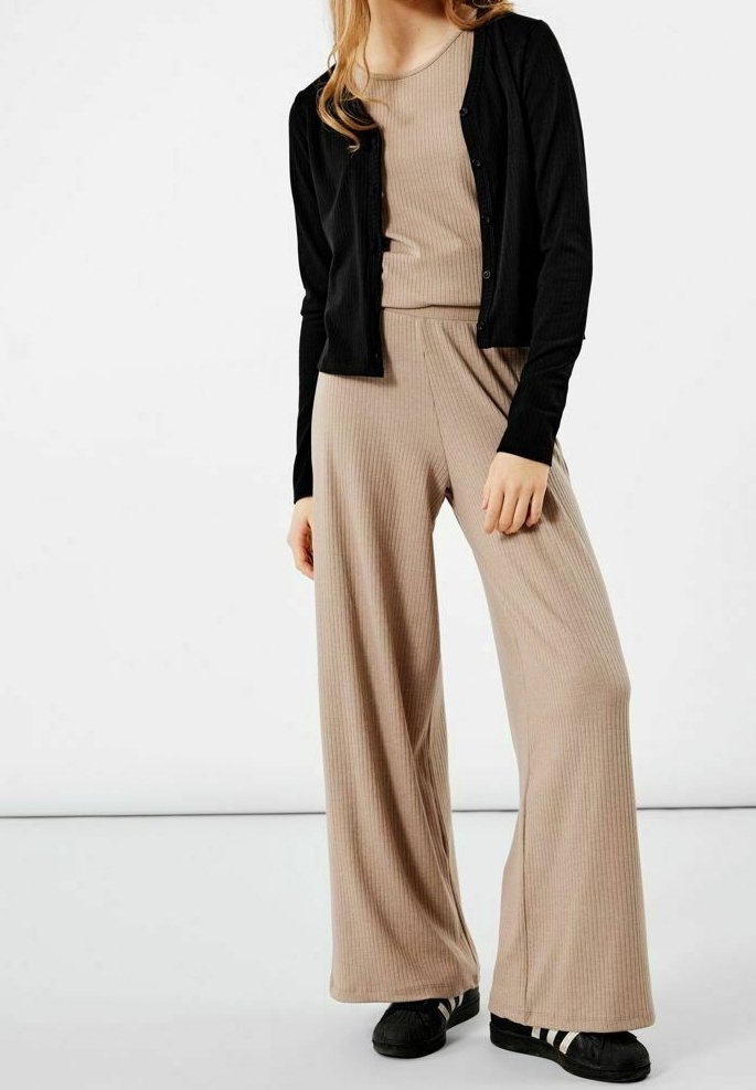 Femme portant un cardigan noir boutonné sur un haut côtelé beige et un pantalon large avec des baskets noires.