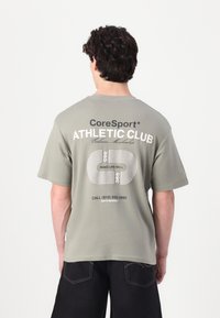 Lys grøn t-shirt med trykt tekst "CoreSport ATHLETIC CLUB" på ryggen. Korte ærmer, rund halsudskæring og afslappet pasform. Mørke bukser synlige.