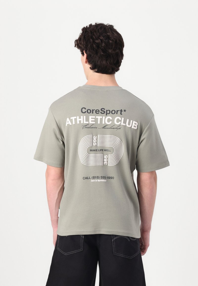 Lys grøn t-shirt med trykt tekst "CoreSport ATHLETIC CLUB" på ryggen. Korte ærmer, rund halsudskæring og afslappet pasform. Mørke bukser synlige.