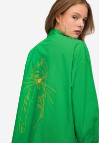 Camicia verde a maniche lunghe con vestibilità rilassata, caratterizzata da un design ricamato giallo di un volto sul retro. Il testo include "Moin Moin." Tessuto dalla consistenza liscia.