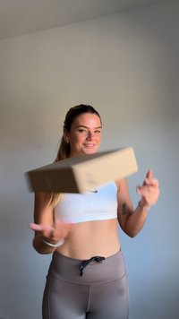 Boîte en carton tenue par une personne, de forme carrée, couleur brun clair. Texture lisse avec un logo sur un côté, en mouvement sur un fond neutre.