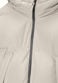 Veste matelassée beige clair avec col montant, fermeture éclair devant et poches latérales montrée en gros plan.