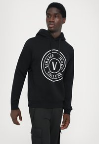 Must harjaga, mis on valmistatud puuvillast, millel on suur valge ümmargune logo, kus on kirjas "VERSACE JEAN COUTURE" ja silmatorkav "V" keskel.