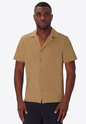 Uomo che indossa una camicia marrone chiaro a maniche corte con bottoni e colletto camp, in piedi contro uno sfondo bianco semplice.