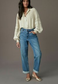 Beige texturerad blus med knappar, högmidjade blå denimjeans och gyllene ballerina-skor. Uppvikta manschetter på jeansen, avslappnad passform överlag.