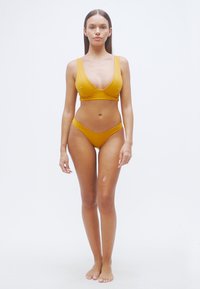 Σετ μπικίνι σε μουσταρδί κίτρινο, με τριγωνικό τοπ και κάτω μέρος χαμηλής μέσης. Λείο ύφασμα με ελάχιστες ραφές και καθαρές γραμμές.